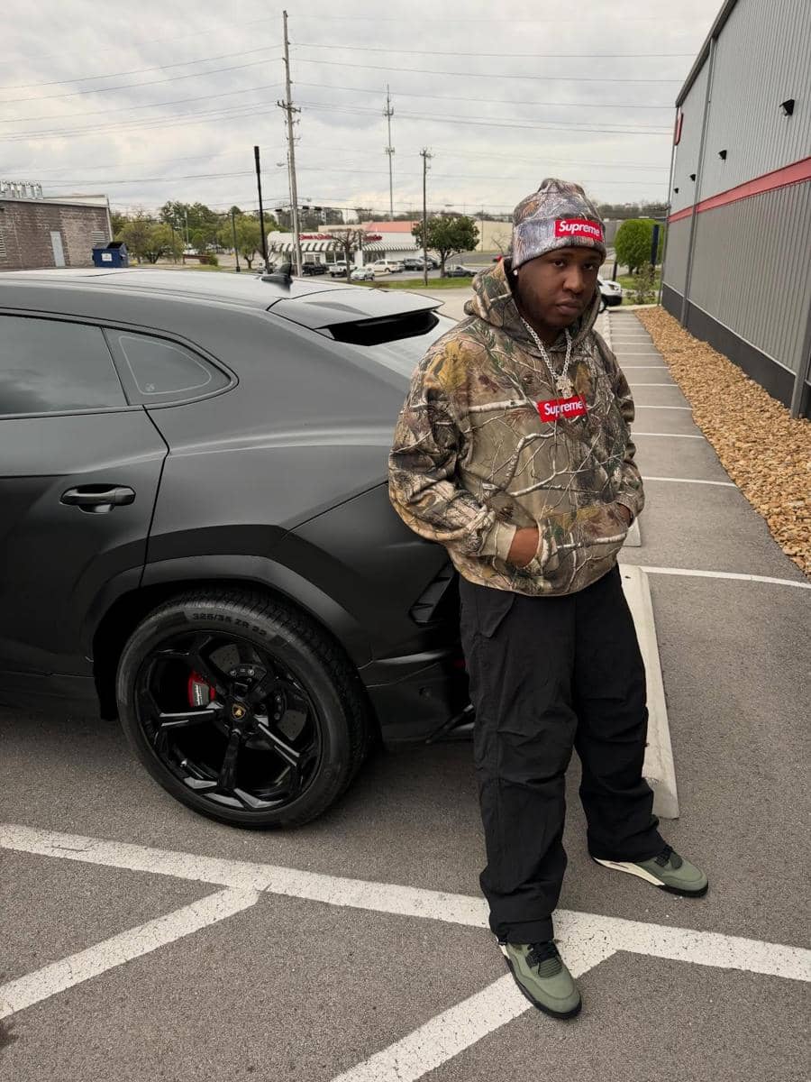 Seorang pria mengenakan hoodie dan topi bermotif kamuflase dengan logo Supreme berdiri di samping mobil sport hitam di area parkir.