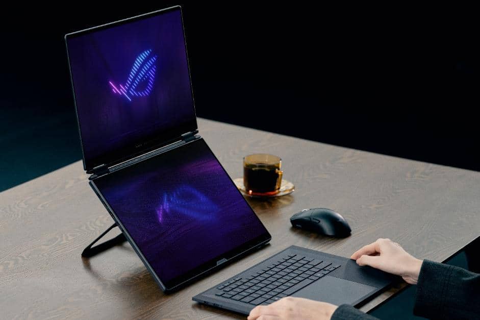 Laptop ROG Zephyrus Duo dengan dua layar terbuka di atas meja kayu, dilengkapi mouse dan secangkir kopi di sampingnya.