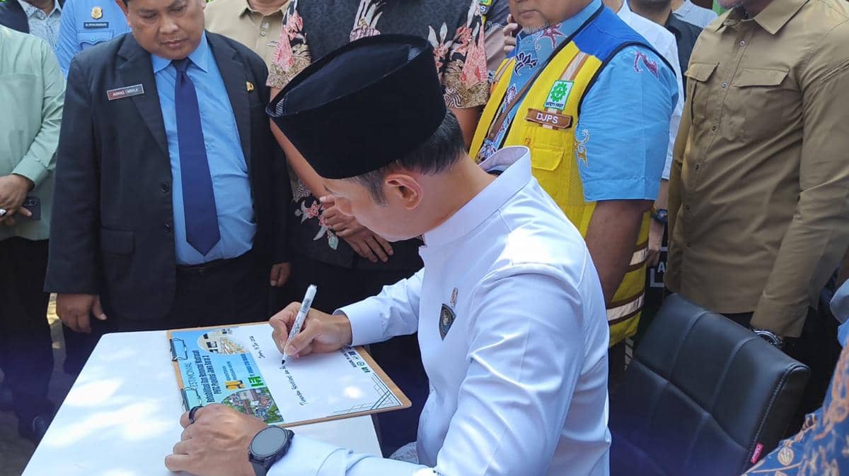 Seorang pejabat mengenakan peci hitam menandatangani dokumen di meja, disaksikan beberapa orang dalam acara resmi di Bogor.
