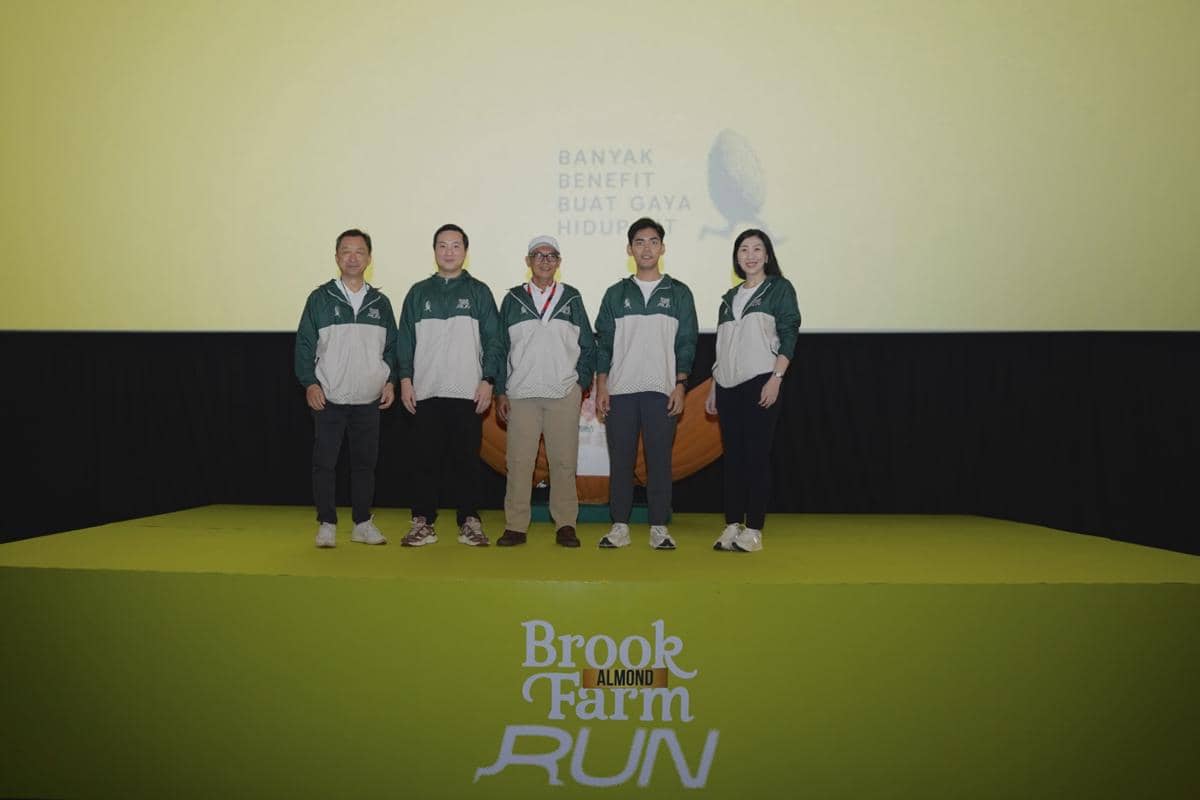 Lima orang mengenakan jaket hijau abu-abu berdiri di atas panggung dengan latar bertuliskan BrookFarm Almond Run 2026.