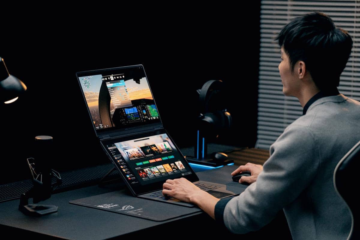 Seseorang menggunakan laptop gaming ROG Zephyrus Duo dengan dua layar di meja kerja modern yang diterangi lampu meja.
