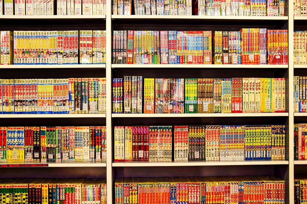 Rak buku berisi berbagai komik dan buku cerita berwarna-warni tersusun rapi di beberapa tingkat rak di sebuah toko atau perpustakaan.