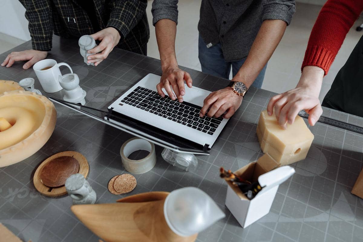Beberapa orang bekerja di meja dengan laptop dan prototipe produk, membahas desain menggunakan bahan dan model fisik.