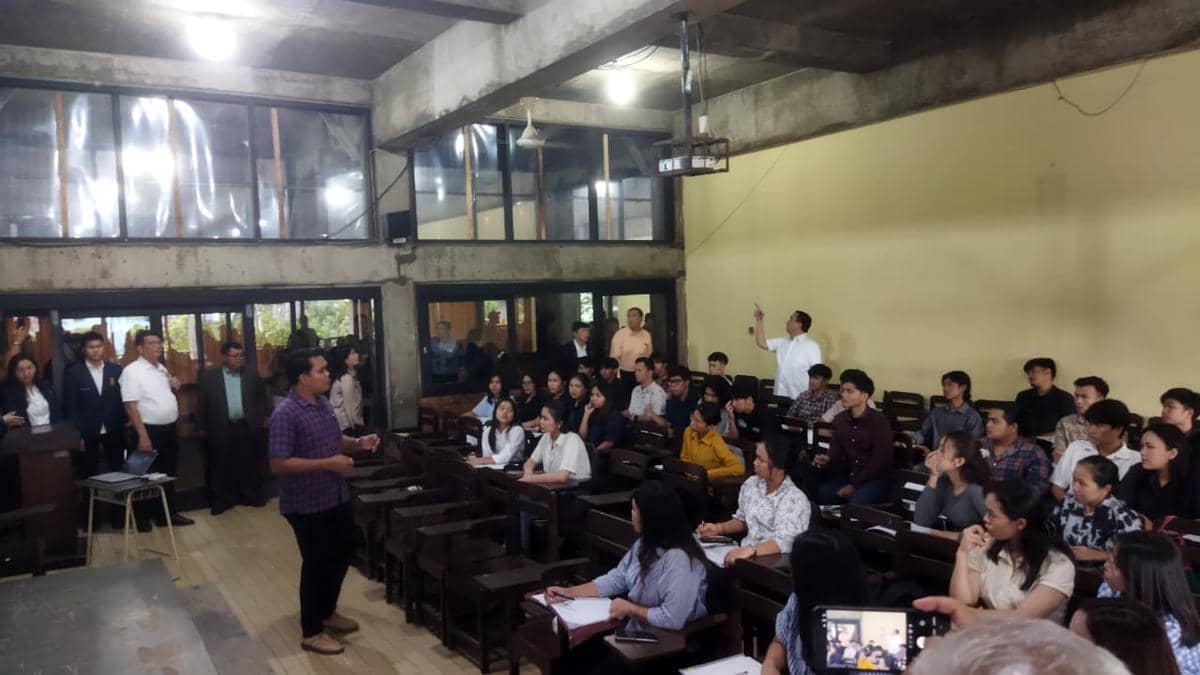 Seorang pria berdiri di depan kelas berbicara kepada mahasiswa yang duduk di bangku kayu, sementara beberapa orang berdiri mengamati di belakang ruangan kampus.
