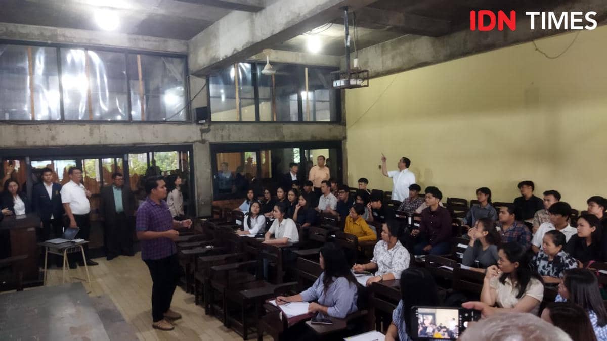 Seorang pria berdiri di depan kelas berbicara kepada mahasiswa yang duduk di bangku kayu, sementara beberapa orang berdiri mengamati di belakang ruangan kampus.