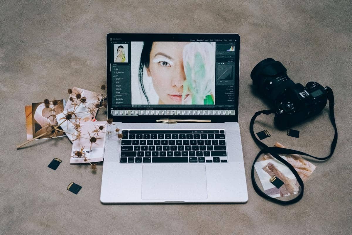 Laptop menampilkan proses pengeditan foto potret di atas meja dengan kamera digital, kartu memori, dan beberapa cetakan foto di sekitarnya.