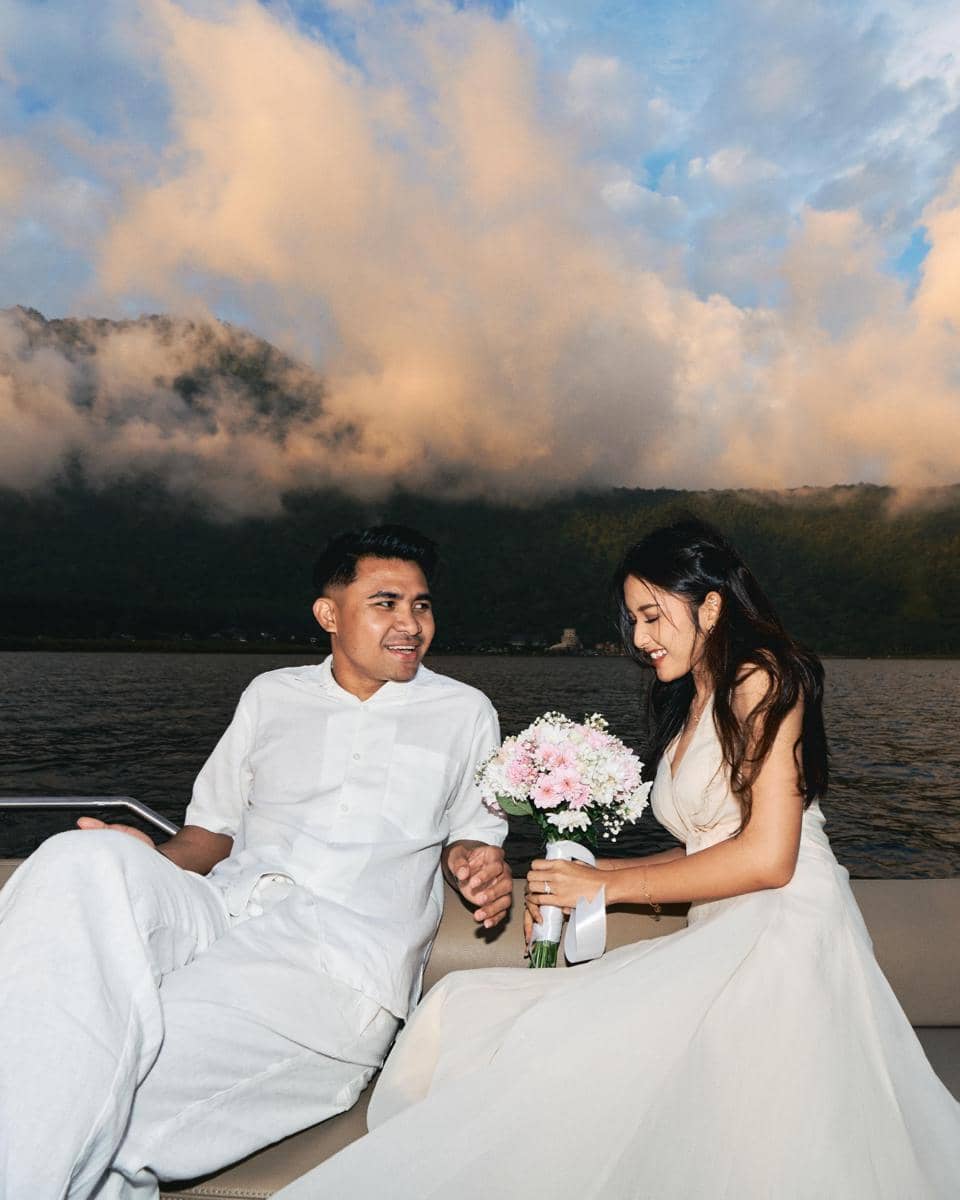 Seorang pria dan wanita berpakaian putih duduk di perahu dengan latar pegunungan dan langit berawan, sambil tersenyum dan memegang buket bunga.