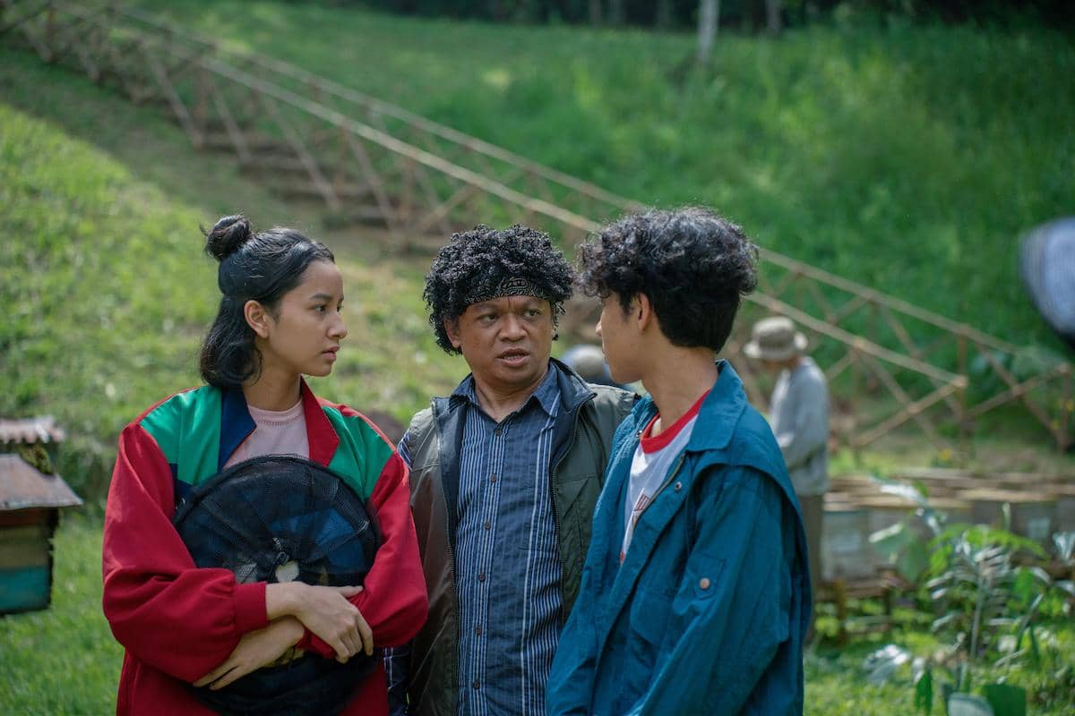 Tiga orang berdiri di area hijau terbuka, dua anak muda berbicara dengan seorang pria paruh baya di tengah mereka, suasana tampak serius.