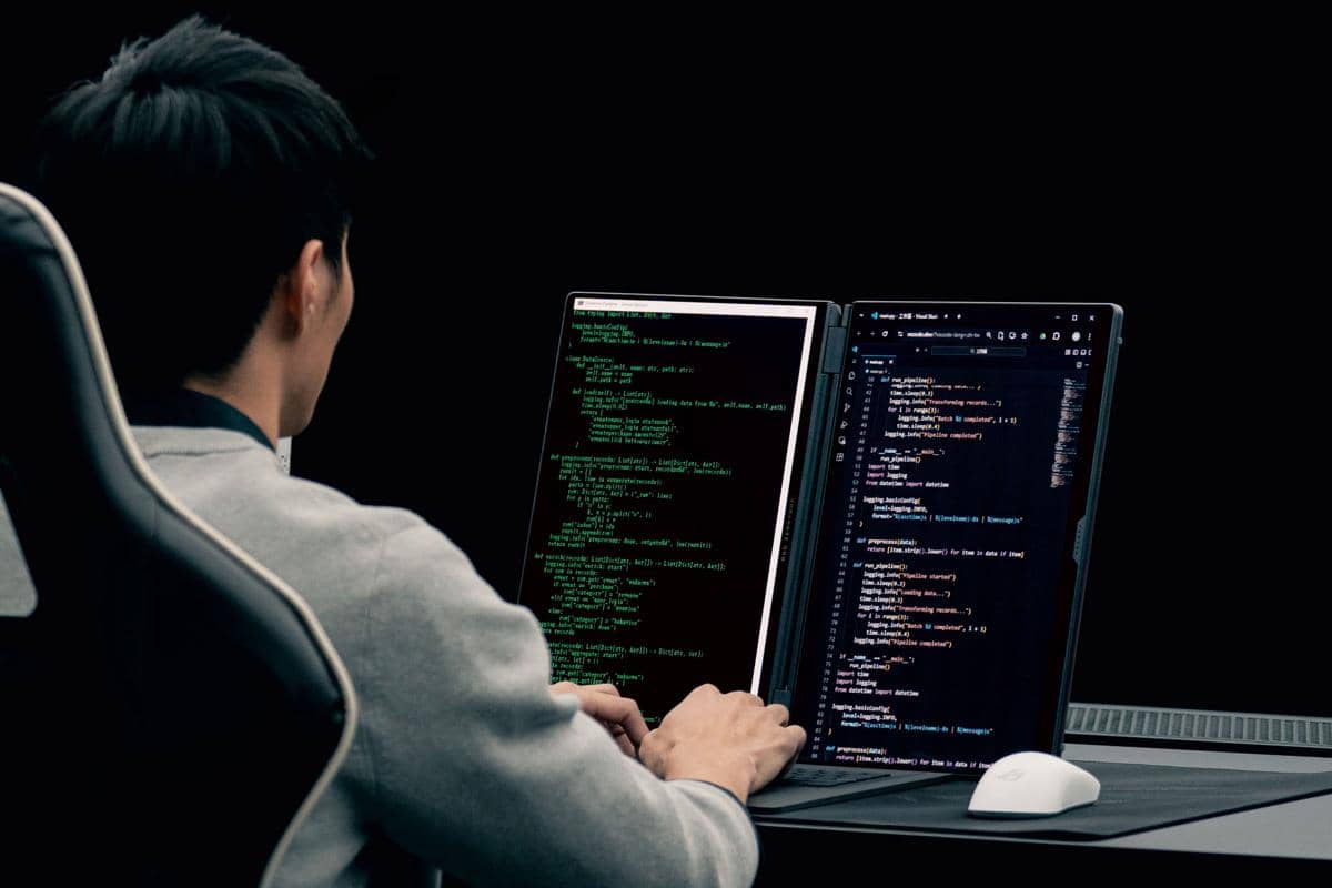 Seorang pengguna duduk di depan laptop ROG Zephyrus Duo dengan dua layar, sedang mengetik kode pemrograman di layar utama.