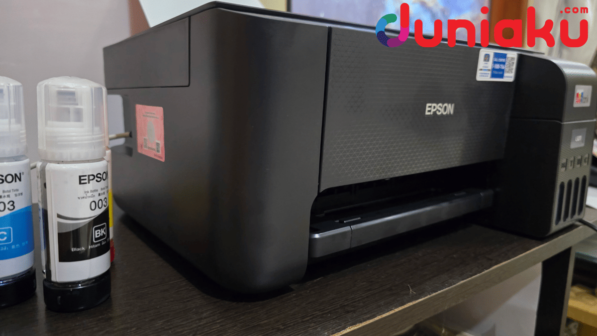 Printer Epson L3211 berwarna hitam diletakkan di atas meja kayu dengan dua botol tinta Epson 003 hitam dan biru di sebelahnya.
