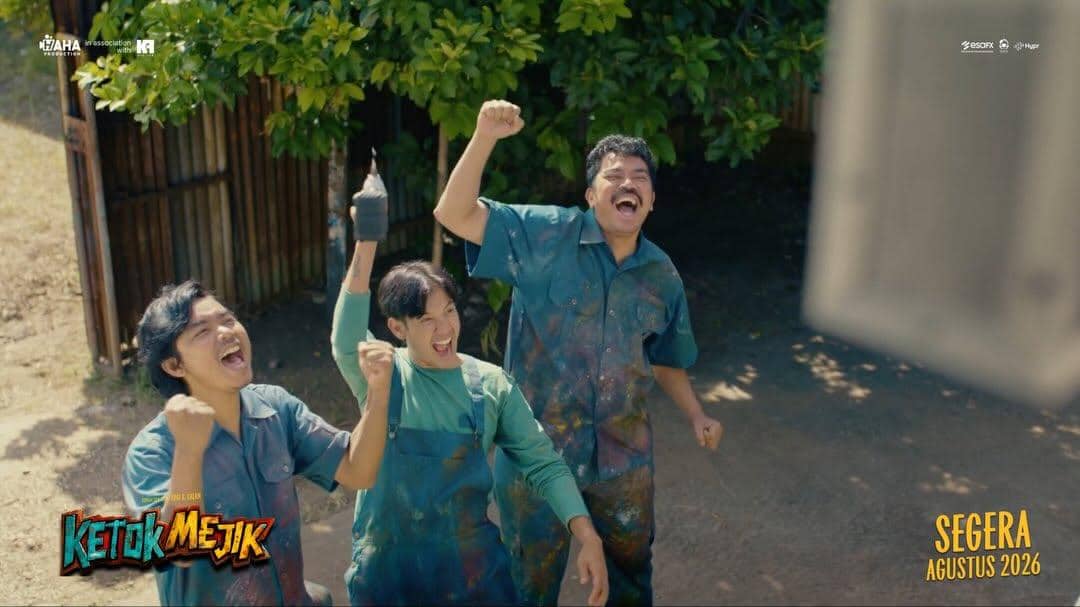 Tiga pria mengenakan pakaian kerja bernoda cat tertawa dan mengangkat tangan dengan ekspresi gembira di luar ruangan.