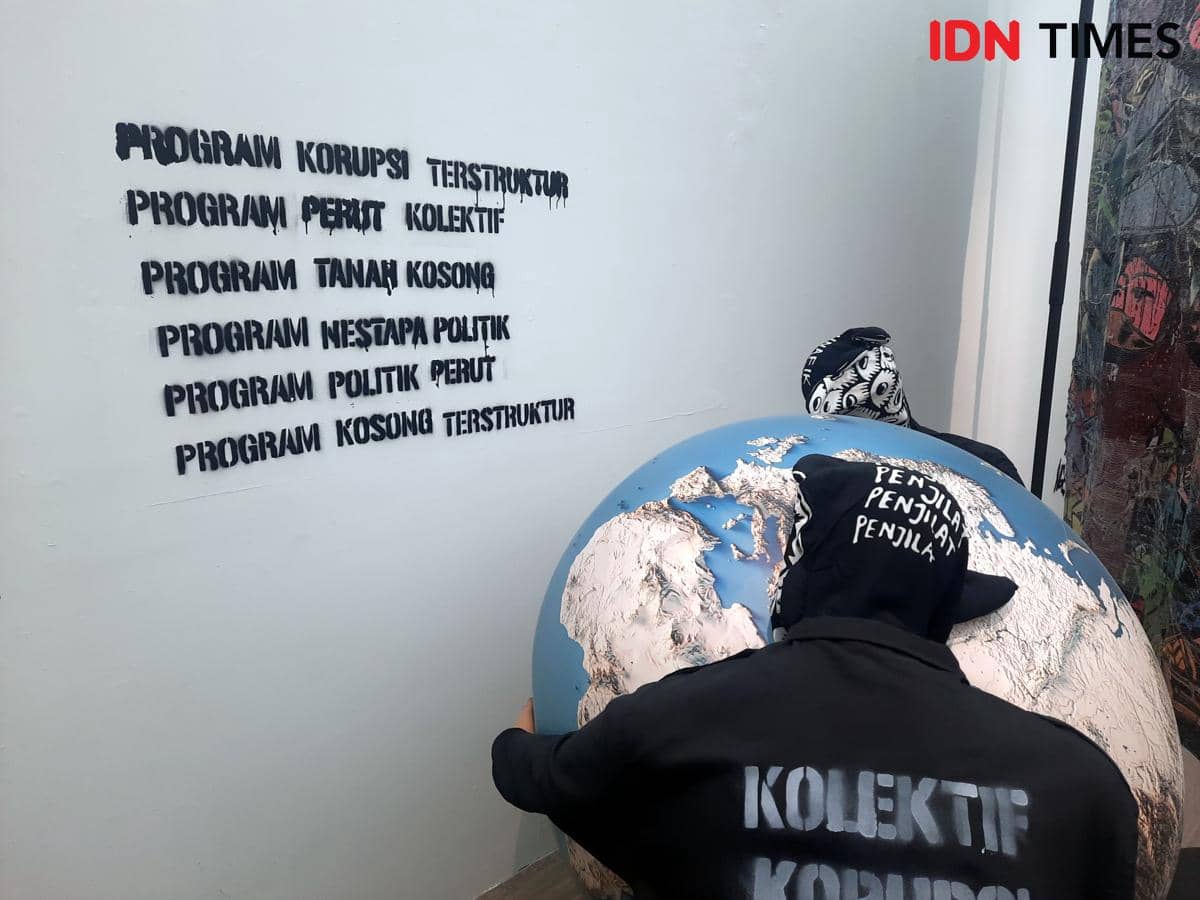 Dua figur mengenakan pakaian hitam bertuliskan pesan kritis memegang bola dunia besar dalam instalasi seni di ARTJOG 2026.