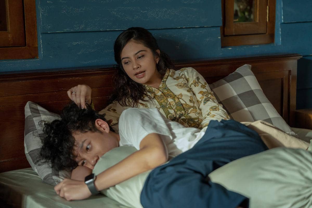 Dua karakter dalam film Aku Sebelum Aku berbaring di tempat tidur, satu terlihat memeluk bantal sementara yang lain berbicara lembut di sampingnya.