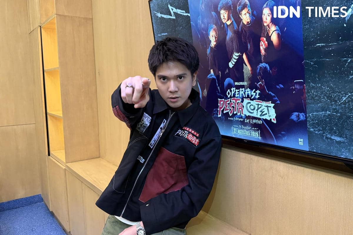 Seorang pria berpose menunjuk ke kamera di depan layar yang menampilkan poster film Operasi Pesta Copet di sebuah ruangan kayu.