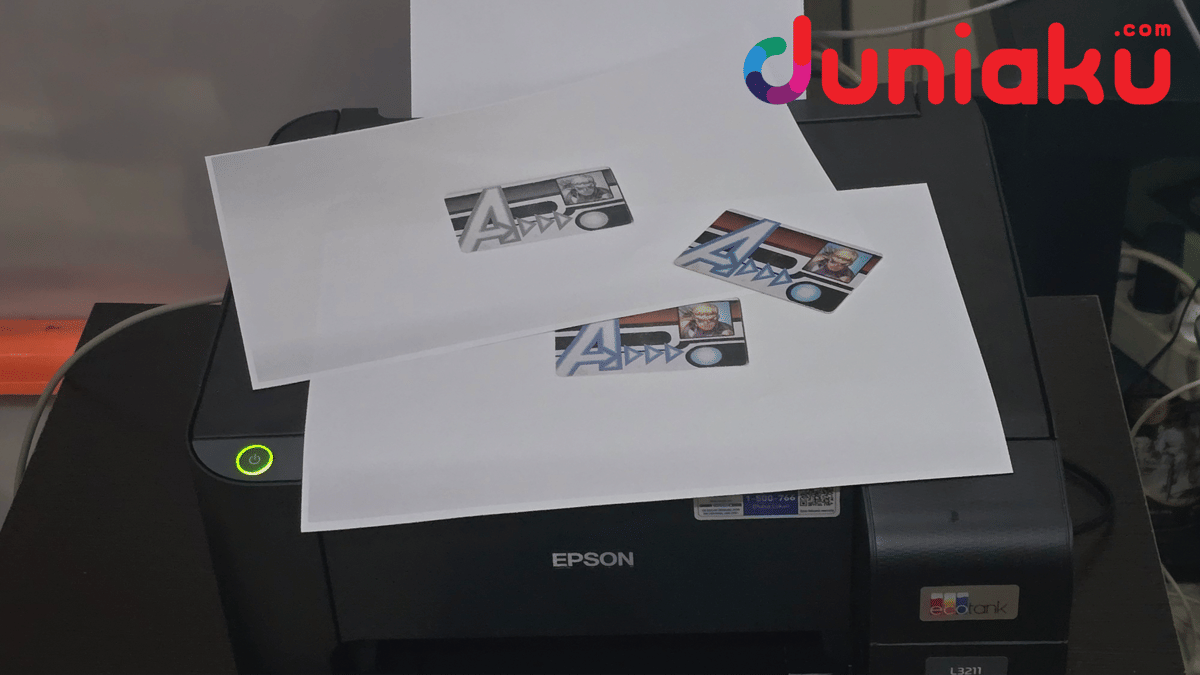 Printer Epson L3211 sedang mencetak hasil fotokopi warna dan hitam putih dengan kertas berisi gambar logo di atasnya.