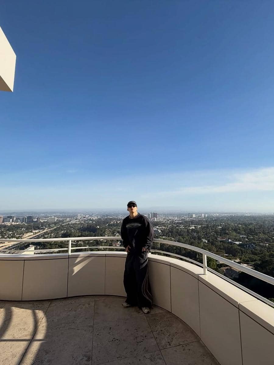 Seorang pria mengenakan sweater hitam, celana longgar, dan topi berdiri di balkon tinggi dengan pemandangan kota di bawah langit biru cerah.