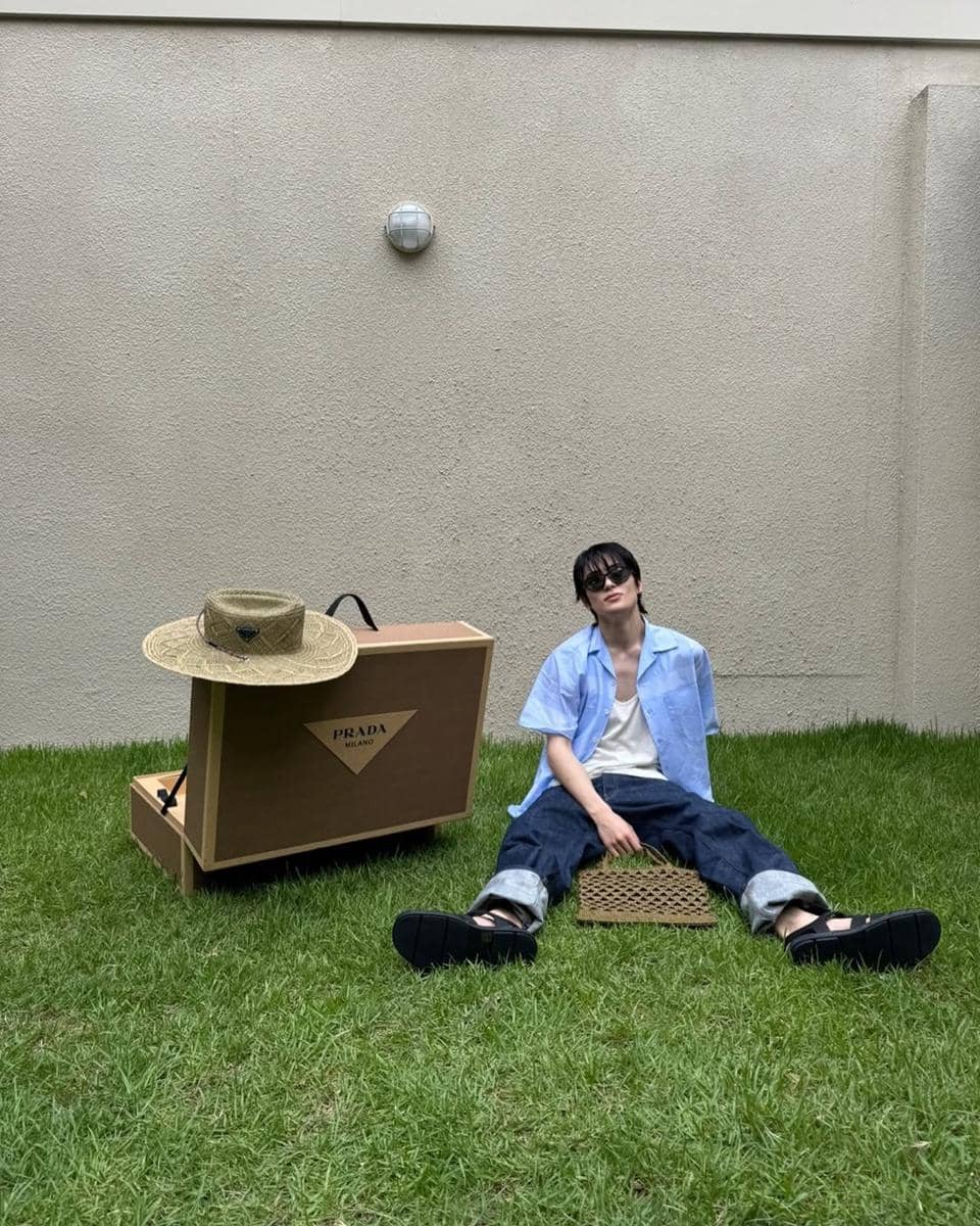 Seorang pria duduk santai di atas rumput mengenakan kemeja biru muda, kaus putih, celana jeans longgar, dan kacamata hitam di samping kotak besar bermerek Prada.