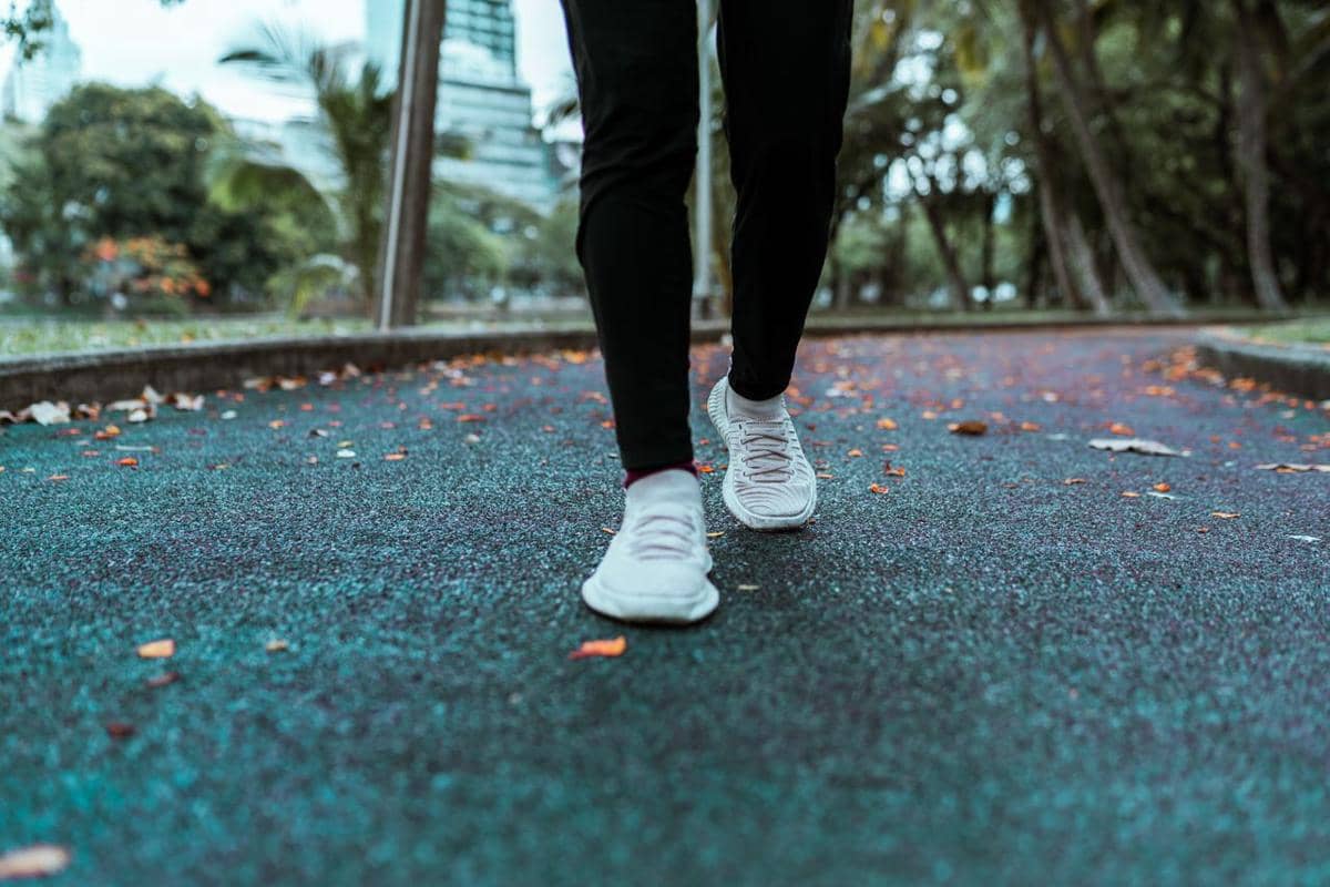 Seseorang berjalan kaki di jalur taman beraspal dengan sepatu olahraga putih dan dedaunan berserakan di sekitarnya.