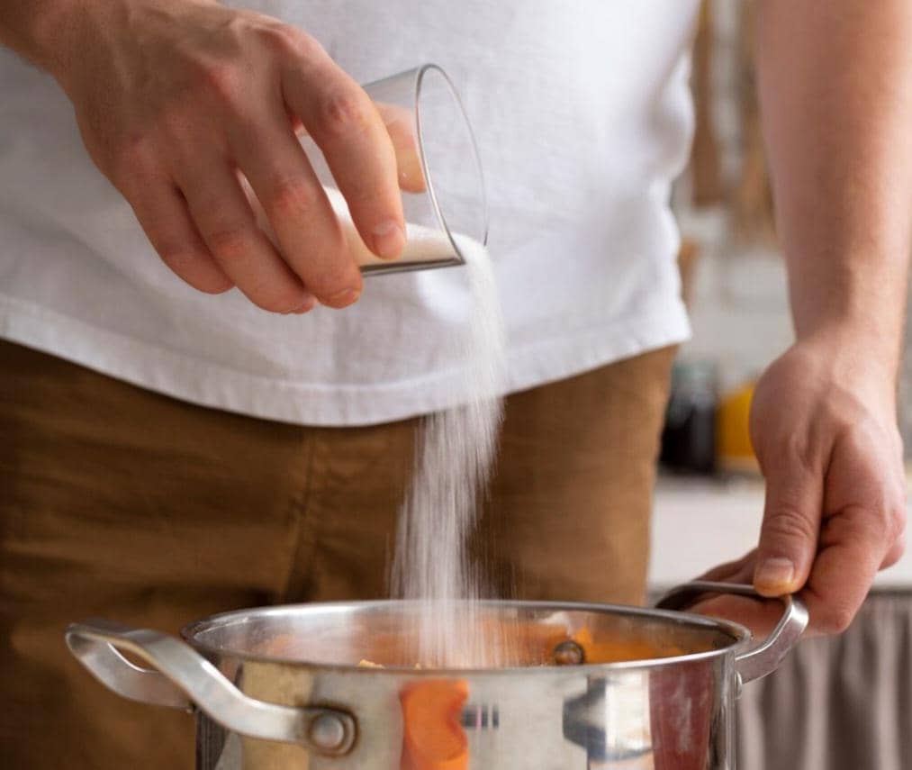 Seseorang menuangkan gula pasir dari gelas ke dalam panci stainless di dapur, mengenakan kaus putih dan celana cokelat.