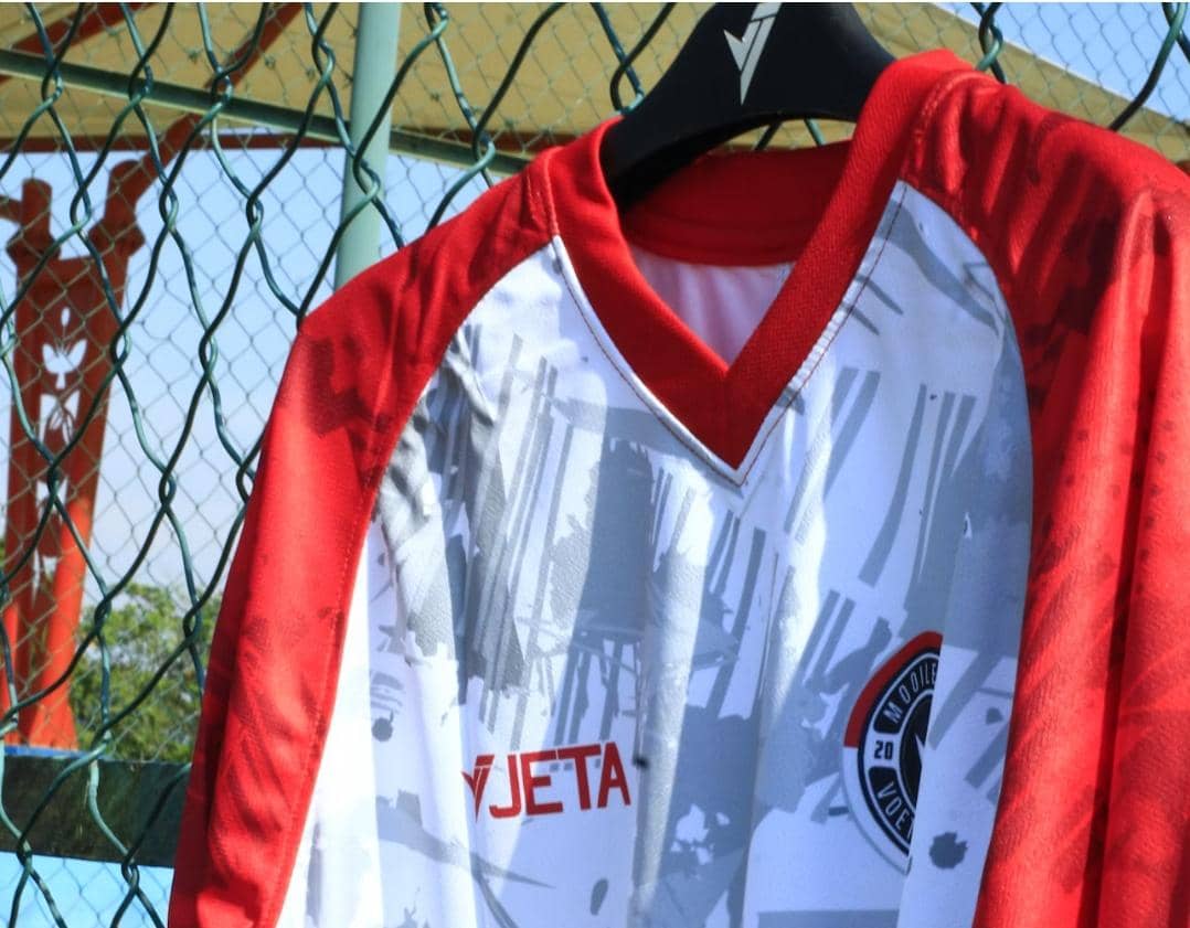 Jersey olahraga berwarna merah dan putih dengan motif abu-abu tergantung di gantungan di dekat pagar kawat lapangan terbuka.