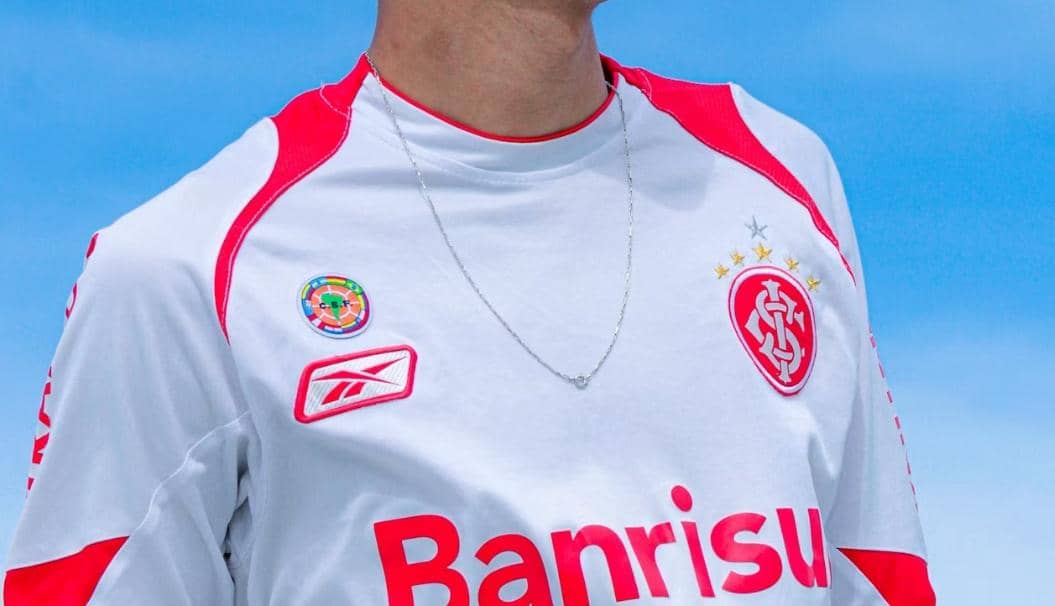 Seseorang mengenakan jersey sepak bola berwarna putih dengan aksen merah dan logo klub di dada di bawah langit biru cerah.