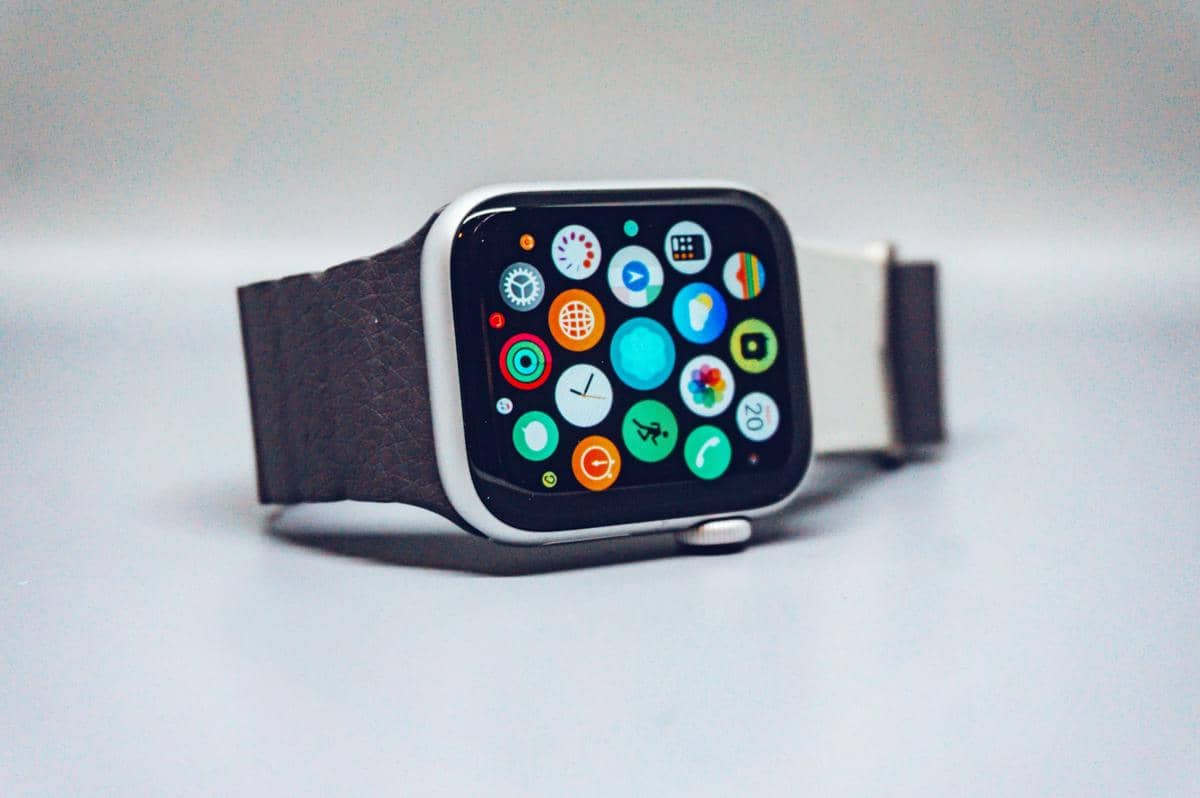 Smartwatch dengan layar berwarna menampilkan berbagai ikon aplikasi dan tali bertekstur kulit berwarna gelap di permukaan putih.