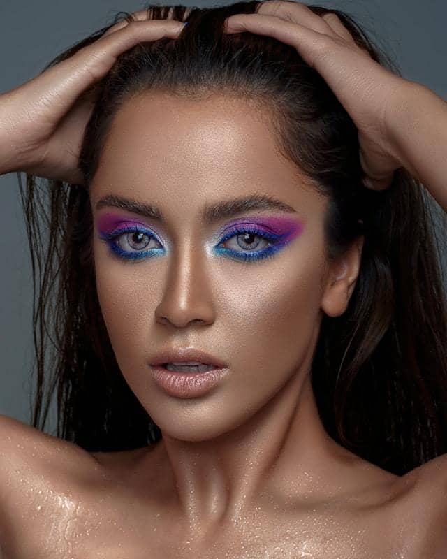 Seorang wanita dengan riasan mata berwarna biru dan ungu mencolok, menatap ke arah kamera dengan tangan di kepala.