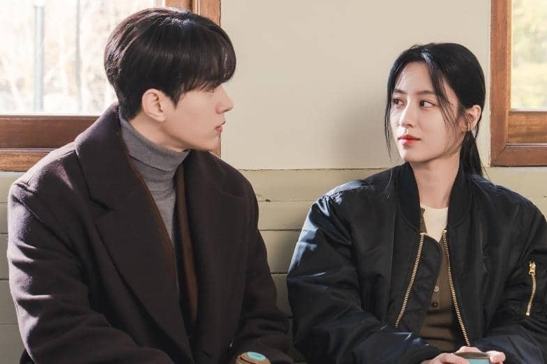 Dua orang duduk berdampingan di dekat jendela, saling menatap dengan ekspresi serius dalam cuplikan drama Love in Sync.