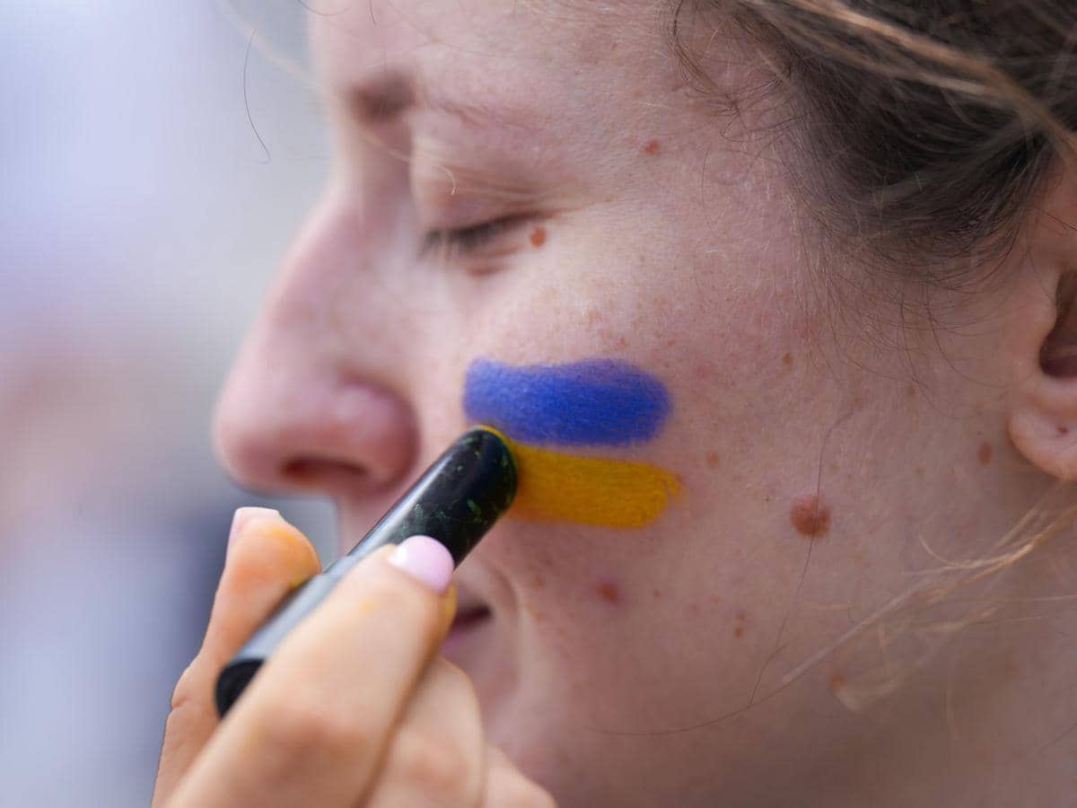 Seorang perempuan menggambar dua garis warna biru dan kuning di pipinya menggunakan facepaint sebagai simbol bendera.