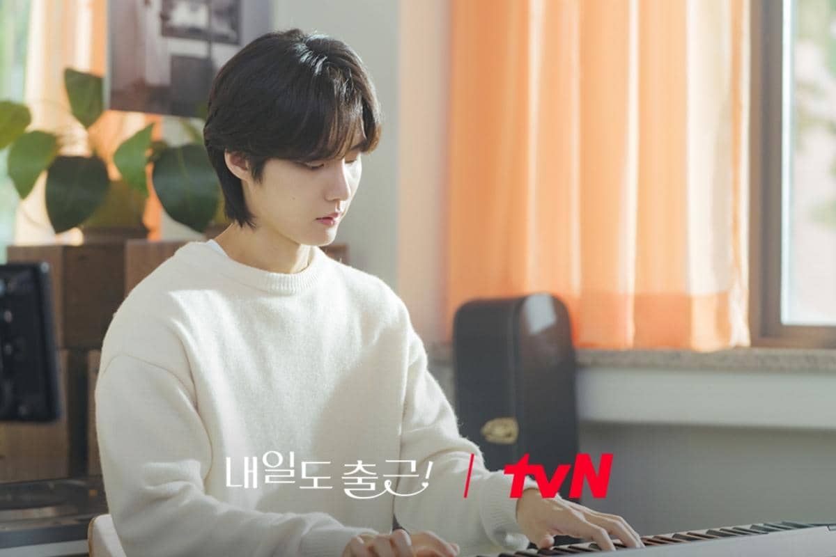 Seseorang mengenakan sweater putih sedang memainkan keyboard di ruangan dengan tirai oranye dan cahaya matahari lembut masuk dari jendela.