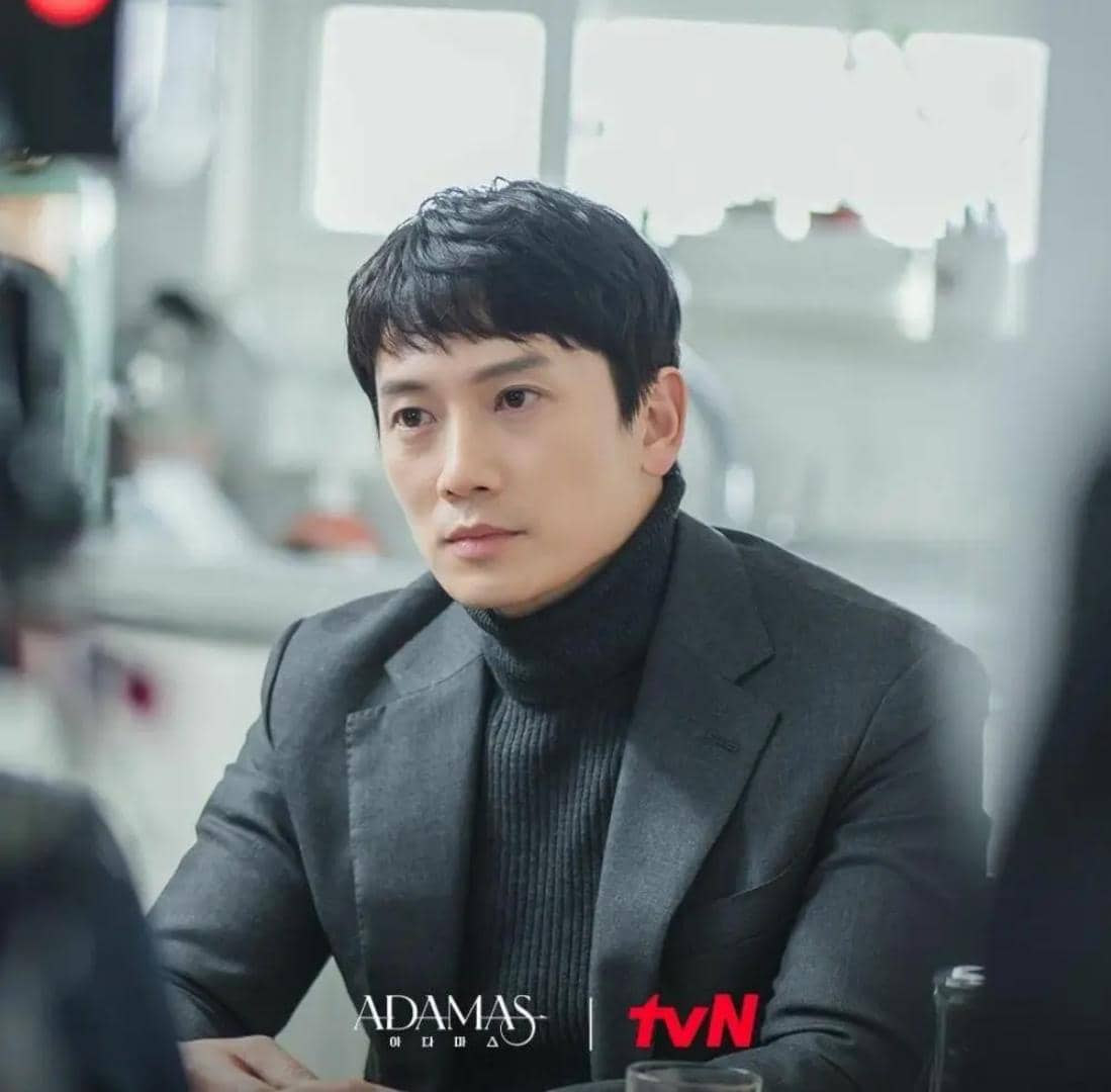 Seorang pria mengenakan setelan jas abu-abu dan turtleneck hitam duduk di dalam ruangan dengan pencahayaan lembut, adegan dari drama Adamas.