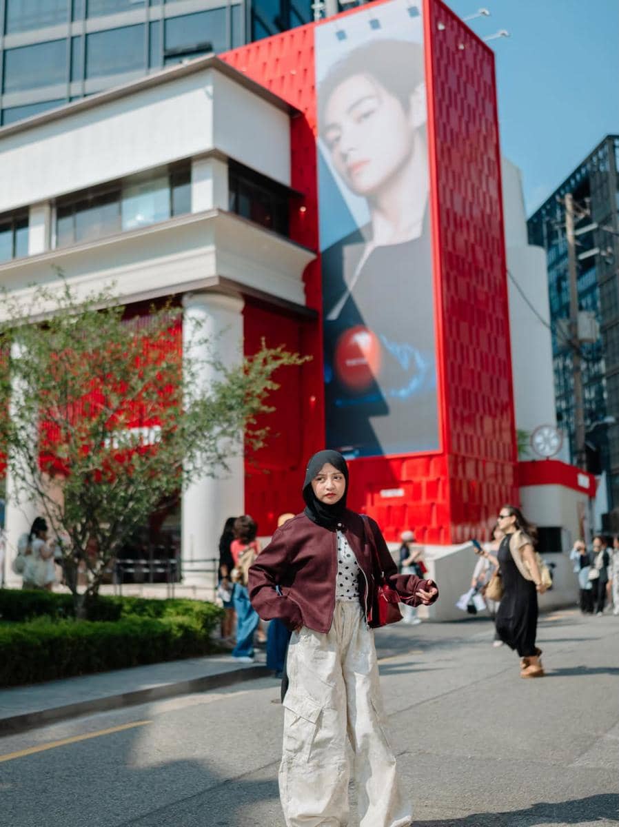 Seorang perempuan berhijab berpose di jalan kawasan Seongsu-dong dengan latar gedung merah dan mural besar di dinding bangunan.