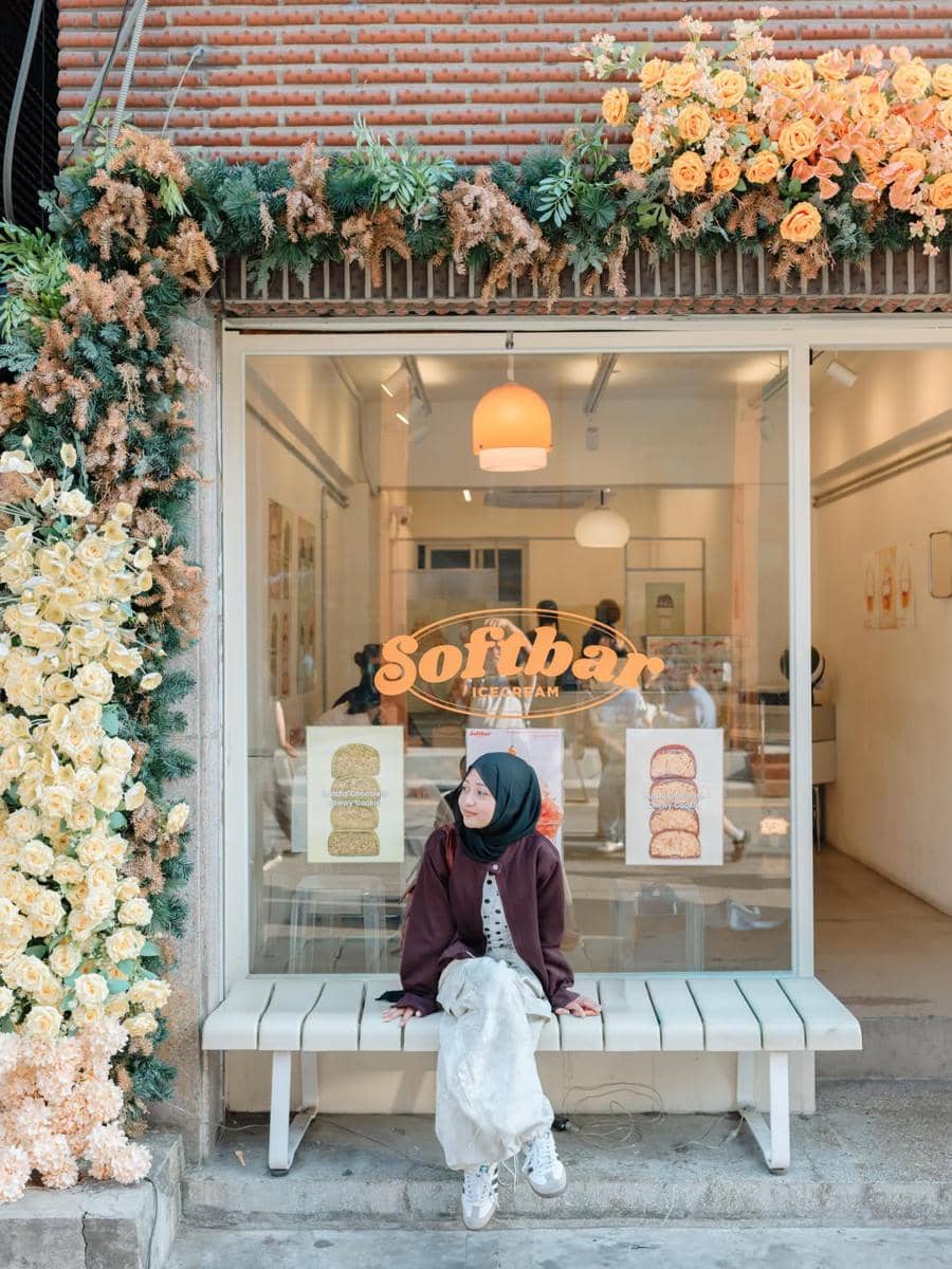 Seorang perempuan berhijab duduk di bangku depan toko es krim Softbar di Seongsu-dong dengan dekorasi bunga berwarna kuning dan hijau.