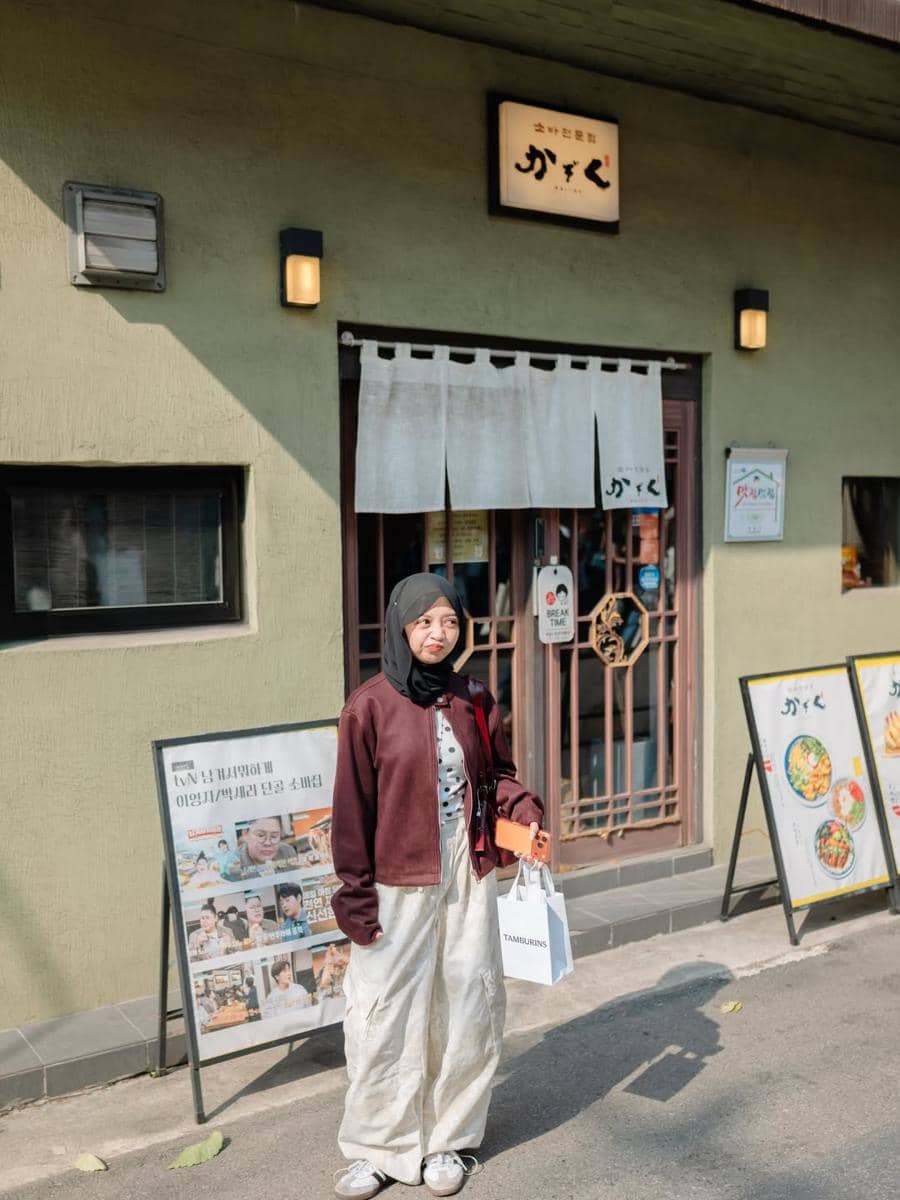 Seorang perempuan berhijab berdiri di depan restoran bergaya Jepang di kawasan Seongsu-dong sambil membawa tas belanja.