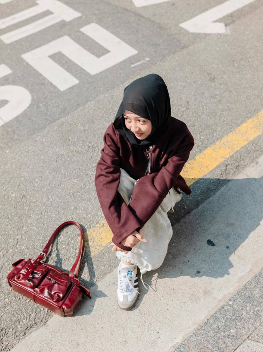 Seorang perempuan berhijab duduk di tepi jalan dengan jaket marun dan tas tangan merah di sampingnya di area Seongsu-dong.