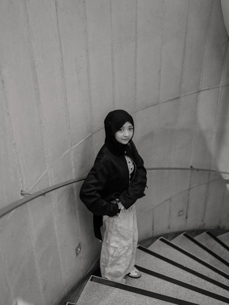 Seorang perempuan berhijab berdiri di tangga melingkar dengan dinding beton di Seongsu-dong, mengenakan jaket gelap dan celana terang.