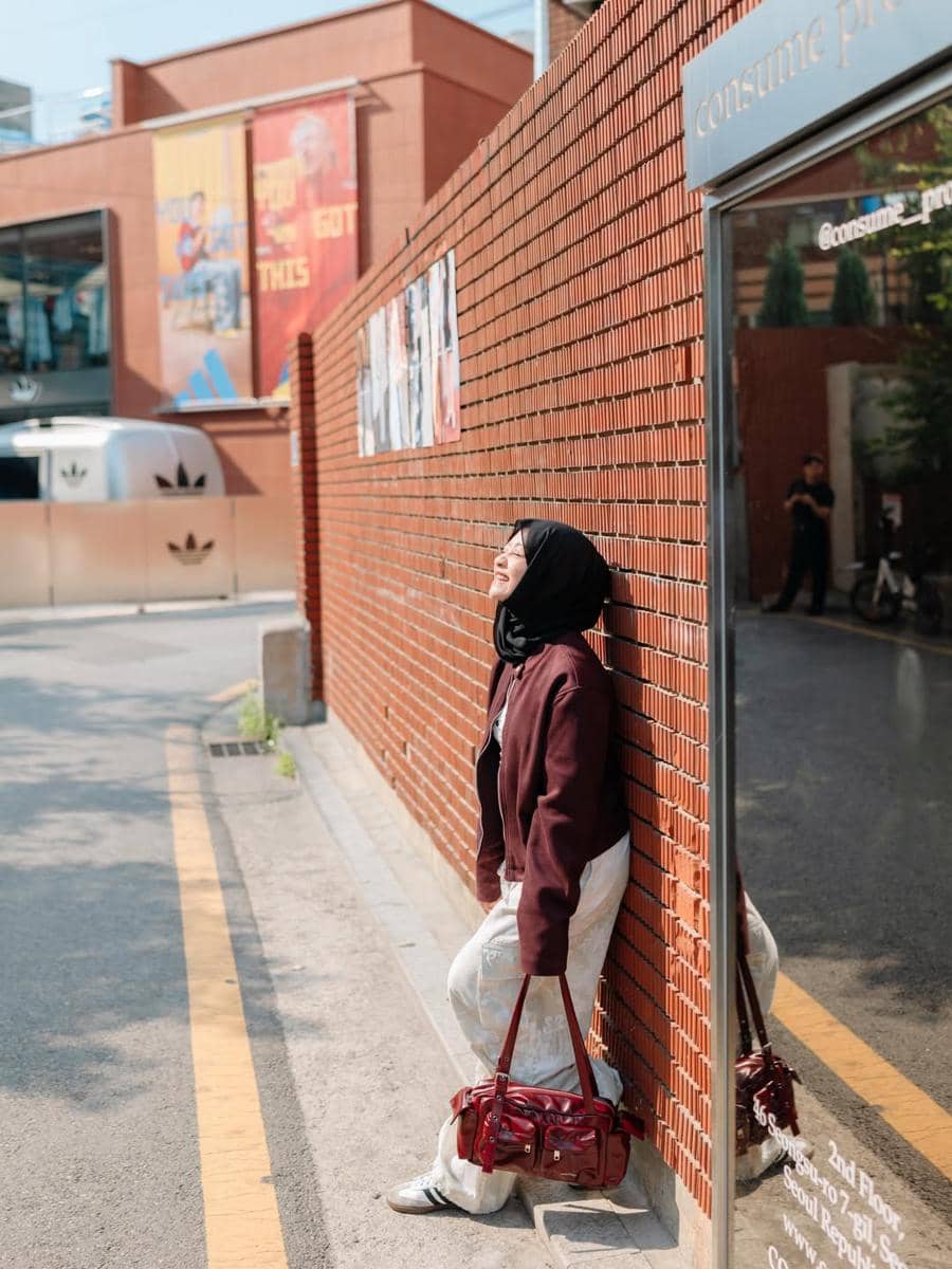 Seorang perempuan berhijab bersandar di dinding bata merah di kawasan Seongsu-dong sambil tersenyum menikmati sinar matahari.