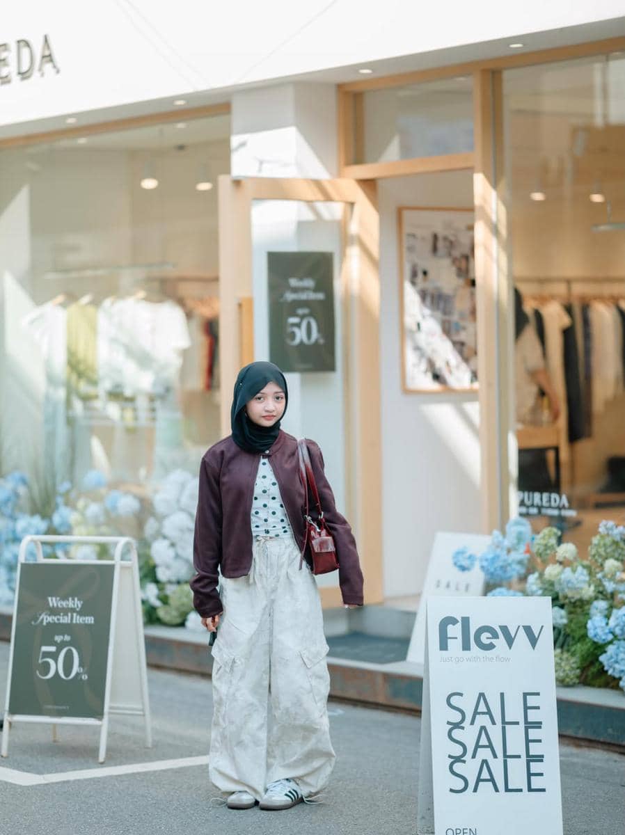 Seorang perempuan berhijab mengenakan jaket marun dan celana putih berdiri di depan toko pakaian dengan papan diskon di Seongsu-dong.