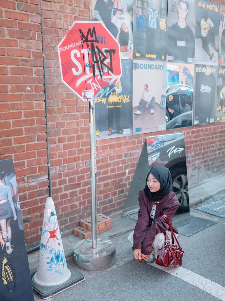 Seorang perempuan berhijab berpose di jalan kawasan Seongsu-dong dengan latar dinding bata dan rambu jalan penuh grafiti.