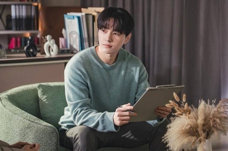 Seorang pria mengenakan sweater biru muda duduk di sofa hijau sambil memegang papan tulis di ruangan dengan pencahayaan hangat.