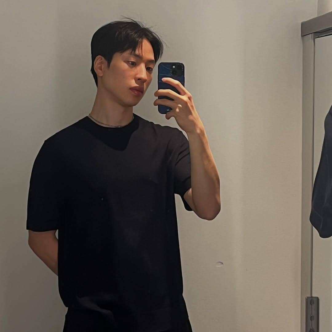 Seorang pria mengenakan kaus hitam berfoto selfie di depan cermin dengan ponsel di tangan kanan di ruangan bercahaya lembut.