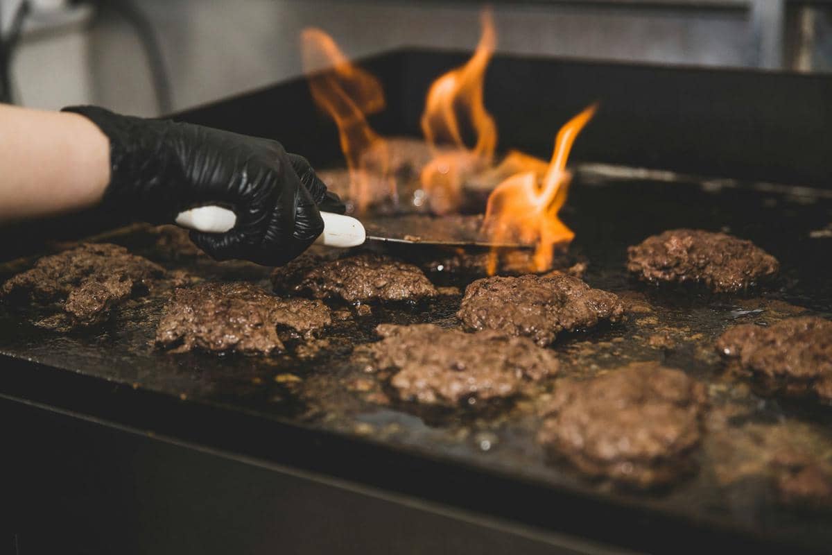 Tangan dengan sarung tangan hitam sedang memanggang beberapa patty daging burger di atas panggangan datar dengan api menyala.