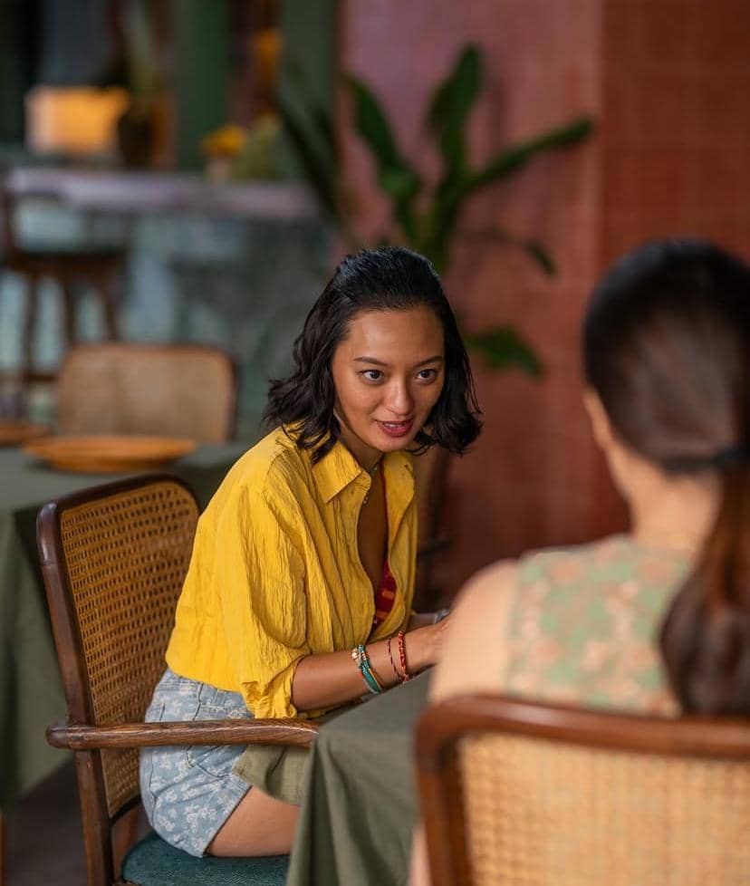 Seorang perempuan berambut pendek mengenakan kemeja kuning dan celana pendek duduk di kursi rotan berbicara dengan seseorang di meja makan.