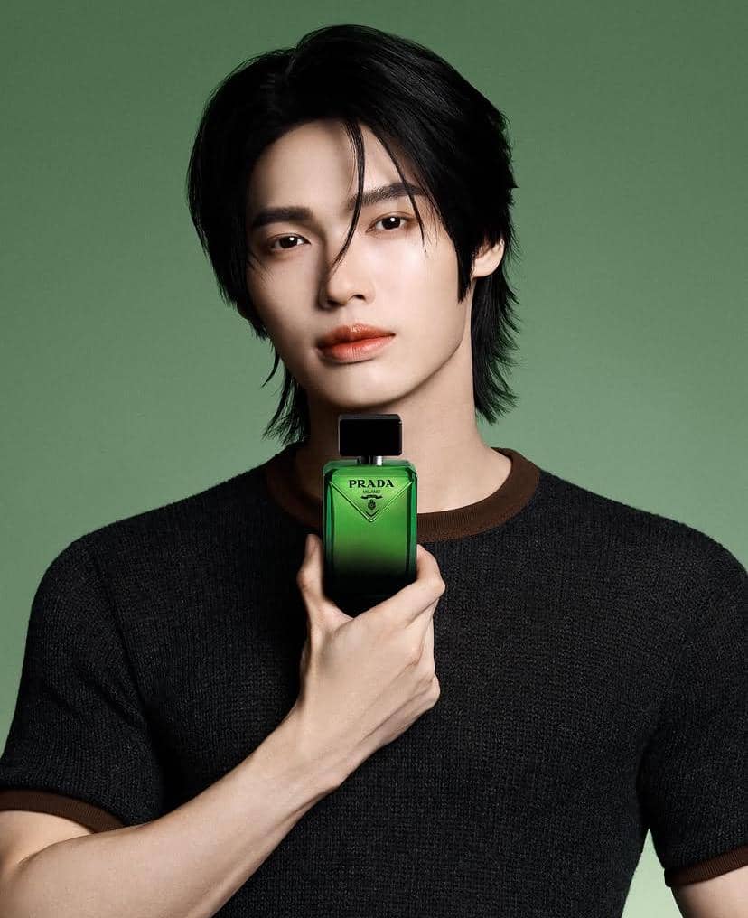 Seorang pria berambut hitam mengenakan kaus gelap memegang botol parfum hijau merek Prada dengan latar belakang hijau.