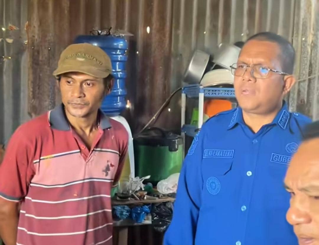 Seorang pejabat daerah mengenakan seragam biru berbincang dengan warga di rumah sederhana berdinding seng dengan peralatan dapur di belakangnya.