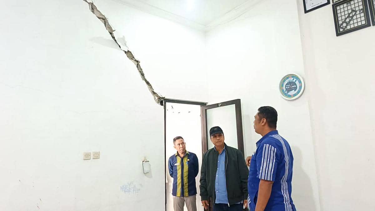 Tiga pria berdiri di dalam ruangan rumah sakit dengan dinding retak besar di atas pintu, meninjau kerusakan bangunan.