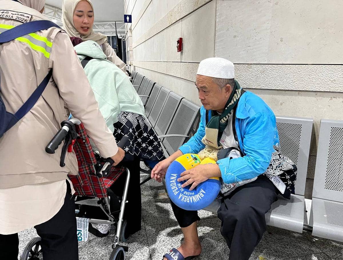 Seorang jemaah haji pria mengenakan kopyah putih duduk di kursi bandara sambil memegang bantal, sementara petugas membantu lansia di kursi roda.