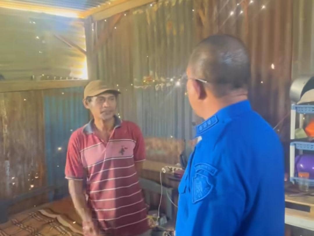 Seorang pejabat berbaju biru berbicara dengan pria berkaus merah di dalam rumah sederhana berdinding seng dan kayu.