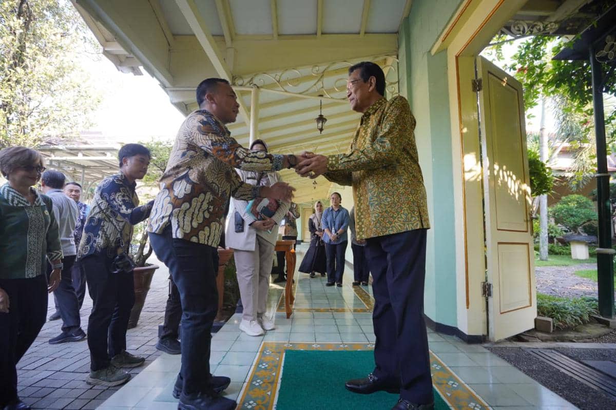 Dua pria berjabat tangan di teras bangunan dengan beberapa orang lain menyaksikan di belakang, mengenakan batik dan pakaian formal.