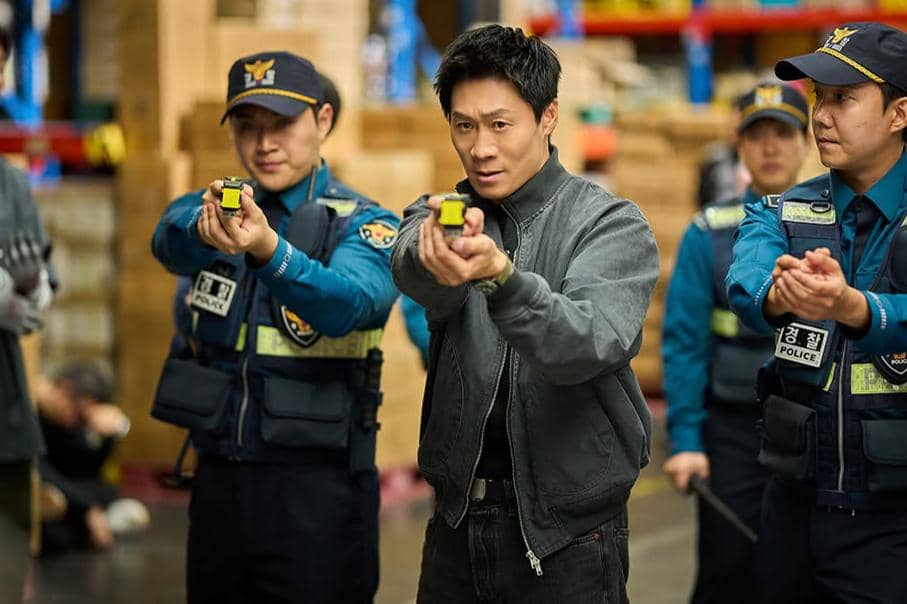 Beberapa polisi mengenakan seragam biru sedang mengarahkan senjata ke depan di sebuah gudang dalam adegan film Husbands in Action.